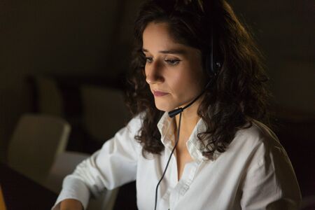 Serious call center operator in dark office. Focused young businesswoman in headset working in dark...