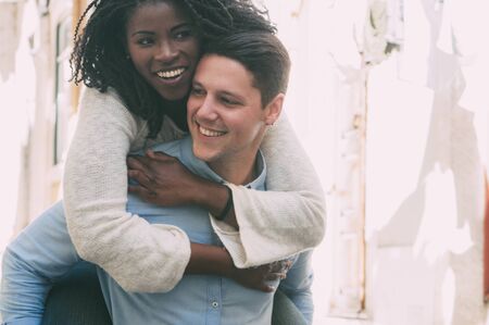Playful guy carrying pretty girlfriend on back outdoors. Happy interracial couple in street. Romance and happiness concept. Front view.の写真素材