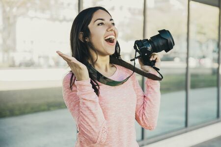 Excited lady taking photos with camera outdoors. Pretty young lady raising hands and standing with mirror glass wall in background. Photography concept. Front view.の写真素材