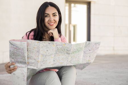 Happy young woman studying paper map outdoors. Pretty lady looking at camera and sitting with building wall in background. Vacation and tourism concept. Front view.の写真素材