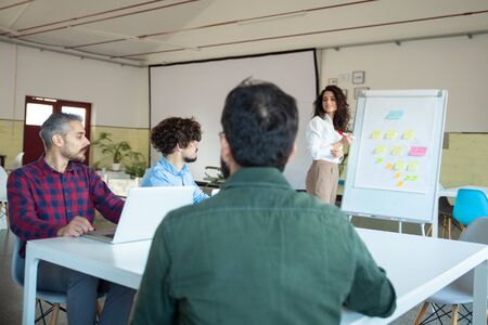 Smiling young woman explaining strategy to coworkers. Confident businesswoman standing near board...