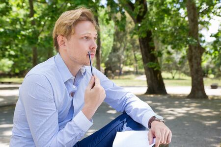 Thoughtful young man sitting in park. Handsome guy holding notepad and pencil. Searching of solution...