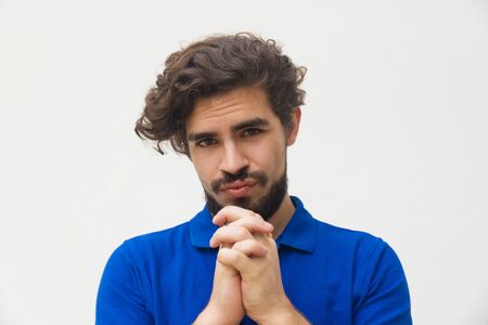 Doubting young man holding hands near chin. Distrustful bearded guy looking at camera. Thinking,...