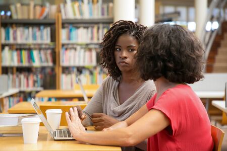 Thoughtful women with laptop at library. Concentrated ladies talking while working with modern device at public library. Education conceptの写真素材