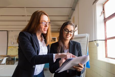 Two smiling women studying new project. Front view of cheerful colleagues reading paper document....