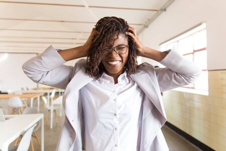 Smiling businesswoman with closed eyes holding hands on head. Emotional African American woman posing in office. Business conceptの写真素材