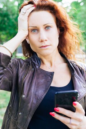 Worried lady holding smartphone and touching head in city park. Pretty young woman looking at camera...