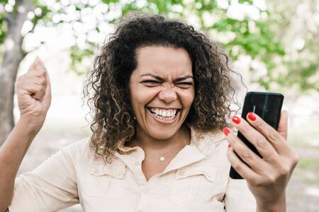 Cheerful woman using smartphone in city park. Young lady pumping fist and standing with blurred green trees and lawn in background. Win concept. Front view.の写真素材