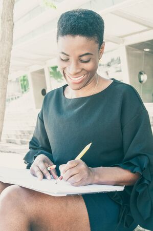 Smiling delighted female artist enjoying leisure time outside. Peaceful African American woman sitting on stairs and drawing with pencils in album. Drawing conceptの写真素材