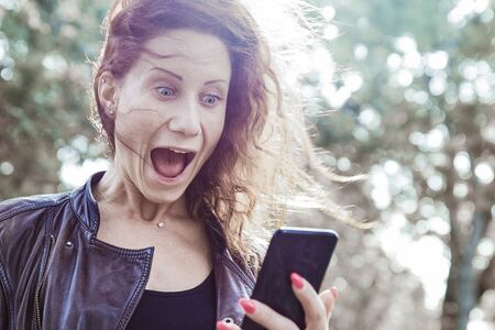 Shocked lady reading news on smartphone in city park. Young woman holding gadget, screaming and standing with blurred green trees in background. Surprising news concept.の写真素材