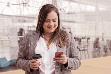 Smiling business woman texting on smartphone outdoors. Lady holding plastic coffee cup, using gadget and standing with building glass wall in background. Break and technology concept. Front view.の写真素材