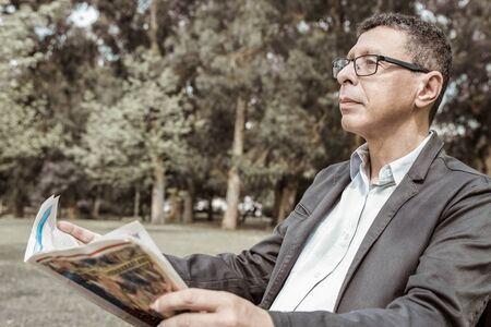 Relaxed man reading newspaper and sitting on bench in park. Guy wearing casual clothes and sitting with green lawn and trees in background. News and nature concept.の写真素材