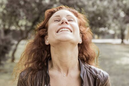 Happy pretty young lady throwing head back in city park. Woman wearing leather jacket and keeping eyes closed with blurred green trees and lawn in background. Happiness concept. Front view.の写真素材
