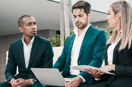 Serious skeptical leader studying report, while managers talking to him. Team of three sitting...