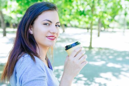 Smiling pretty young woman drinking coffee in park. Beautiful lady wearing hoodie, holding plastic...