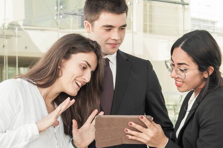 Three good looking serious colleagues standing in office corridor, attractive woman in spectacles showing data on tablet, they looking interested. Work, communication conceptの写真素材