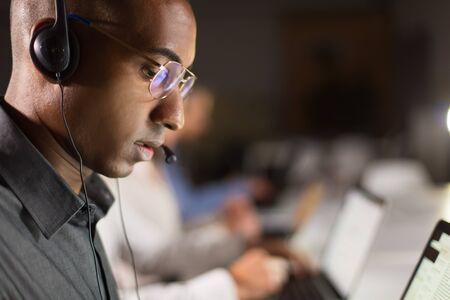 Side view of call center operator typing on laptop. African American young man in eyeglasses working...