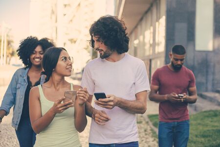 Beautiful young couple with smartphones excited with discussion outdoors. Mix raced man with mobile phone and black woman walking behind them. Communication outdoors conceptの写真素材