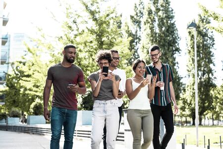 Cheerful friends using their phones for group video calls. Five people walking outside and using two...