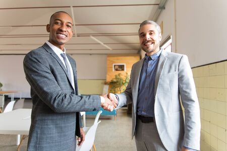 Two smiling businessmen shaking hands and looking at camera. Cheerful young men wearing formal wear posing in office. Business conceptの写真素材