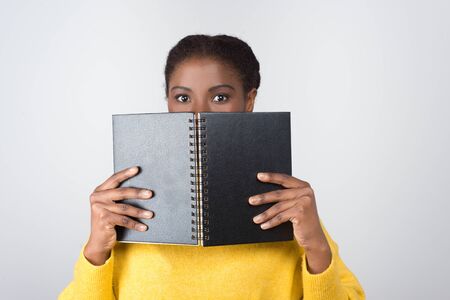 Cute African American student covering face with open notebook. Attractive young lady with notepad looking at camera. Education, tuition conceptの写真素材