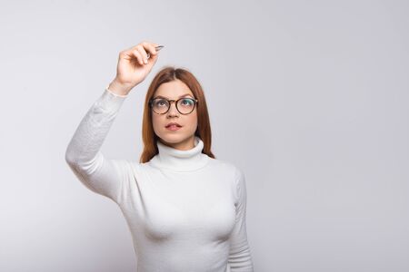 Focused woman writing something in air. Concentrated young woman in eyeglasses holding pen and writing isolated on white background. Business conceptの写真素材