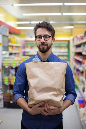 Man holding paper bag and smiling at camera. Handsome happy young man holding grocery bag and looking at camera in supermarket. Shopping conceptの写真素材