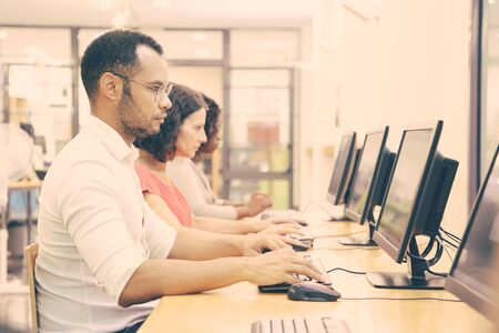 Diverse group of students taking online tests in computer class. Line of man and women in casual...