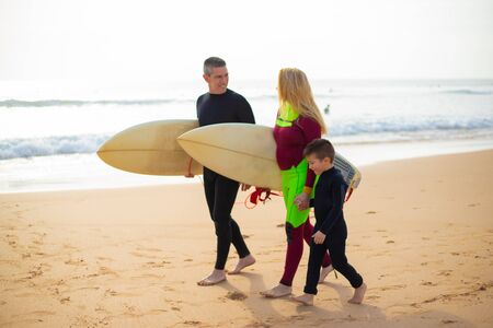 Family with surfboards walking on ocean coast. Side view of happy parents and little son in wetsuits holding hands and walking together on sandy beach. Surfing conceptの写真素材