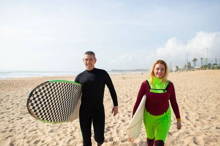 Happy couple of surfers on beach. Front view of cheerful man and woman in wetsuits holding...