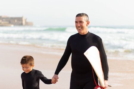Happy father and son with surfboard on beach. Father and son in wetsuits holding hands and walking...