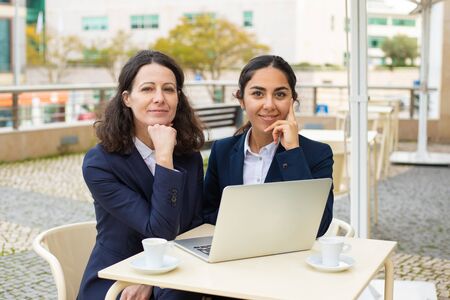 Businesswomen smiling at camera in outdoor cafe. Cheerful female colleagues sitting at table,...
