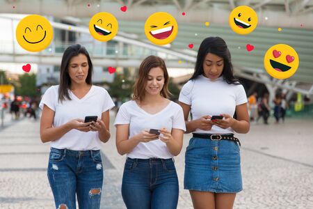 Cheerful friends standing on street and texting on phones with chat icons in background. Front view of smiling young women holding modern smartphones. Technology conceptの写真素材
