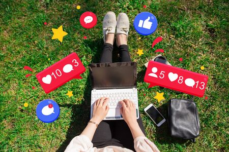 Woman working on laptop in park. Top view of girl sitting on grass with portable pc on her knees and social media icons around, coffee cup, book and smartphone. Working in park conceptの写真素材