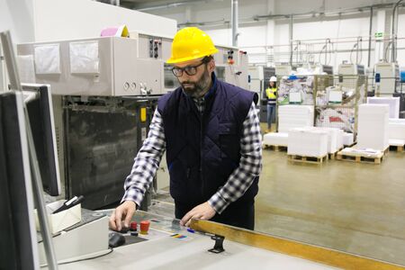 Focused male factory worker operating machine at control panel. Bearded middle aged man in hardhat...