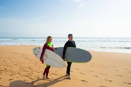 Couple of surfers walking on beach. Side view of middle aged man and woman in wetsuits holding...