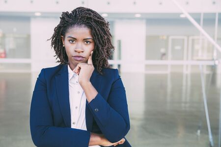Serious pensive professional posing near office building. Young black business woman standing at outdoor glass wall, leaning chin on hand, looking at camera. Business portrait conceptの写真素材
