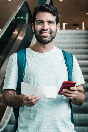 Smiling young man holding ticket and smartphone. Cheerful traveler looking at camera. Travel concept