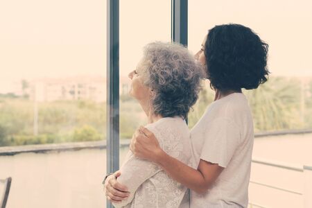 Peaceful middle aged and senior women watching scene from window. Adult daughter embracing old mother at home. Family relationship conceptの写真素材