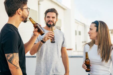 Positive friends enjoying friendly talk and drinking beer on outdoor terrace. Young men and woman in casual meeting outside. Friendly conversation conceptの写真素材
