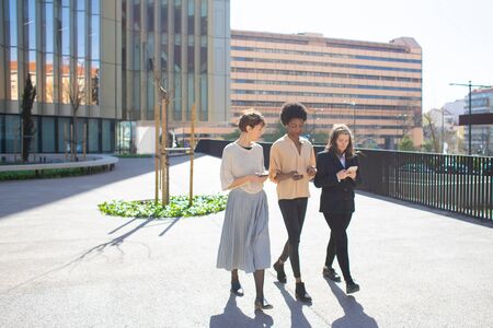 Beautiful young women talking while walking on street. Tranquil ladies strolling with smartphones and having pleasant talk. Technology, communication conceptの写真素材