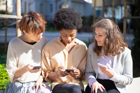 Three women with smartphones sitting on street. Cheerful female friends looking at phone....
