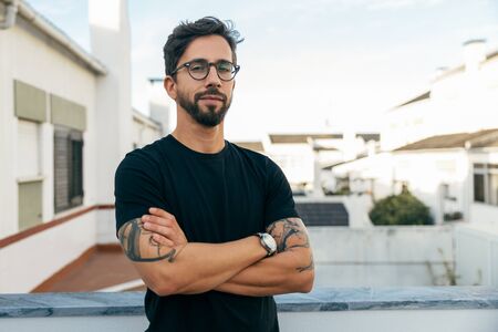Confident stylish guy with tattoos posing on apartment balcony or terrace. Young man in glasses standing outside with arms crossed and looking at camera. Male portrait conceptの写真素材