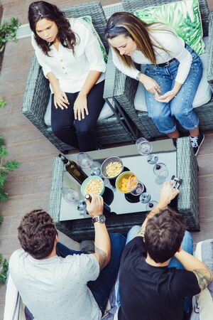 Four friends chatting on terrace over glass of wine. Young men and women in casual meeting outside....