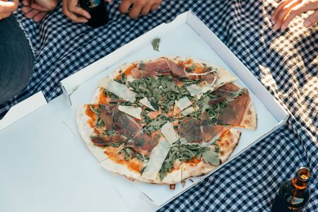 Group of friends eating pizza outdoors. Top view of pizza in box on plaid, beer bottles and human hands. Picnic conceptの写真素材