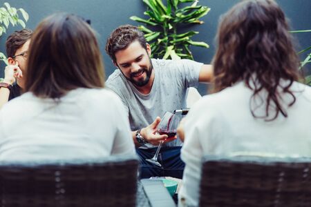 Happy positive bearded guy pouring wine for his friends on outdoor terrace. Young men and women in casual meeting outside. Relaxing and party with friends conceptの写真素材