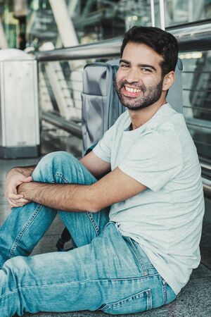 Smiling Hispanic man sitting on floor at airport. Cheerful young guy looking at camera while resting...
