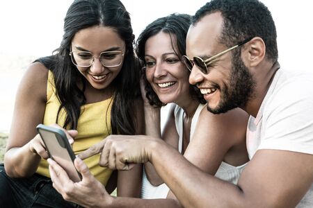 Happy multiethnic friends using smartphone. Smiling young man and women sitting together and using mobile phone in park. Technology conceptの写真素材