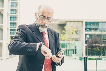 Focused grey haired businessman with cellphone consulting watch on his way to office. Elderly man in...
