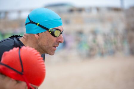 Swimmers in wetsuits on beach. Side view of male and female athletes in sportswear looking aside...